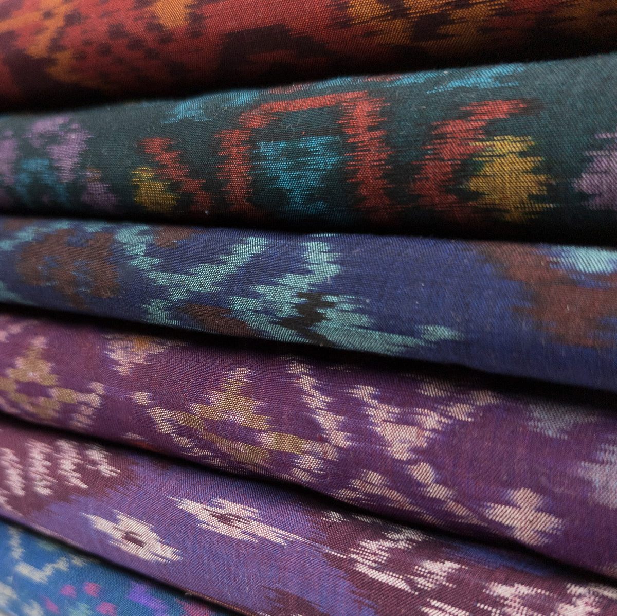 A close up of six bolts of colourful handwoven ikat fabric stacked on top of each other. The fabrics are a mixture of blue, purple, green and red, and each feature a different traditional ikat pattern in contrasting colours.