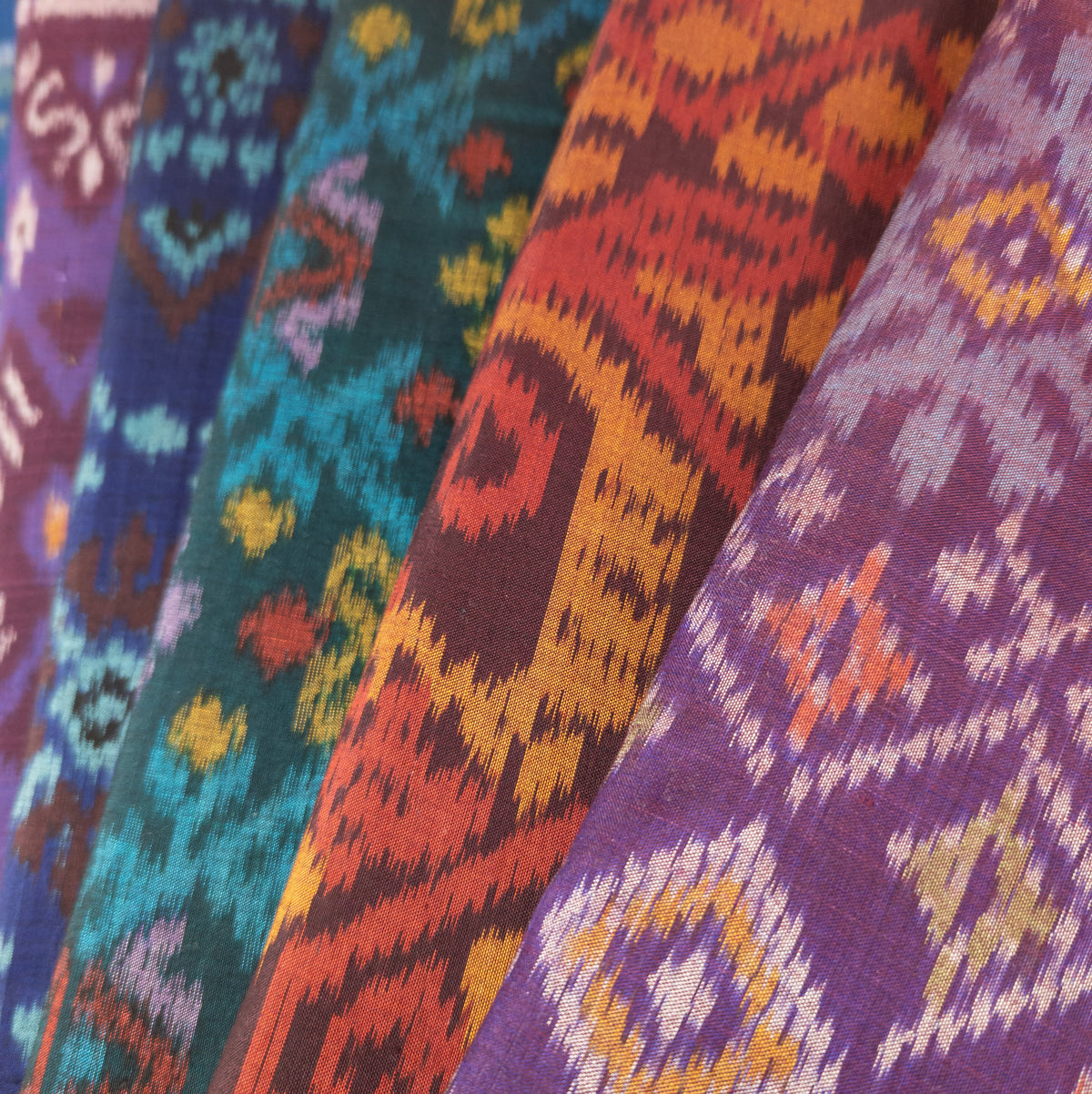 A spread of five different colourful Indonesian ikat fabrics in purple, red, green and blue. Each bolt of fabric has a different traditional woven pattern on it.