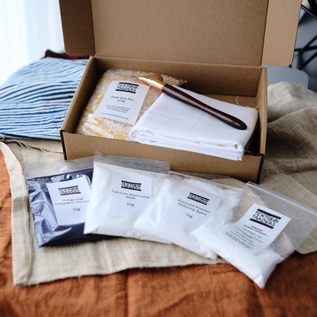 An open cardboard box showing the contents of an indigo batik kit - white folded primissima fabric, a copper tjanting, indigo dye and other powders in packets.