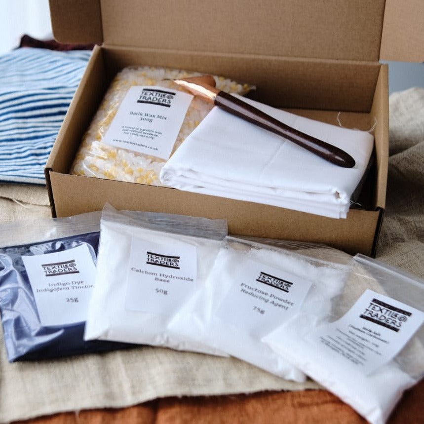 An open cardboard box showing the contents of an indigo batik kit - white folded primissima fabric, a copper tjanting, indigo dye and other powders in packets.