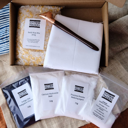 An open cardboard box containing the contents of an indigo batik kit: white folded fabric with a wooden-handled tjanting resting on top, alongside several small labeled powder packets laid out in front on a textured fabric surface.