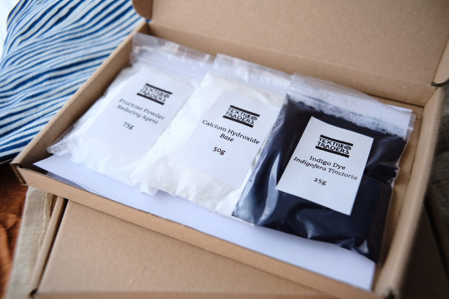 An open kraft cardboard box containing three labeled bags of pigment powders: a white bag labeled 'Fructose Powder Reducing Agent 75g', a white bag labeled 'Calcium Hydroxide Base 50g', and a blue bag labeled 'Indigo Dye Indigofera Tinctoria 25g'.