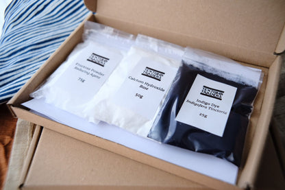 An open kraft cardboard box containing three labeled bags of pigment powders: a white bag labeled 'Fructose Powder Reducing Agent 75g', a white bag labeled 'Calcium Hydroxide Base 50g', and a blue bag labeled 'Indigo Dye Indigofera Tinctoria 25g'.