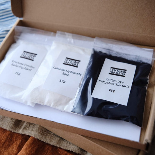 An open cardboard box containing small bags of indigo dye, fructose, and calcium hydroxide placed on a wooden surface with a striped textile nearby.