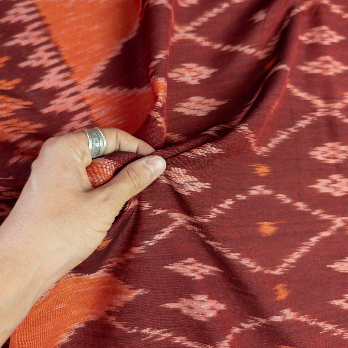 A close-up of a hand holding and draping a rust-red and orange patterned fabric with a coral geometric ikat design.