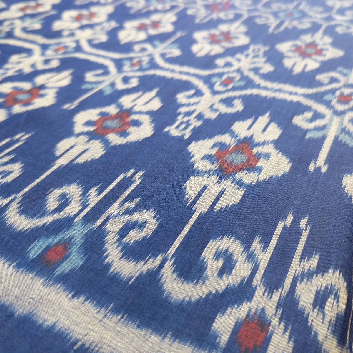 A close-up of a blue fabric with a traditional ikat floral pattern in cream, light blue, and red.