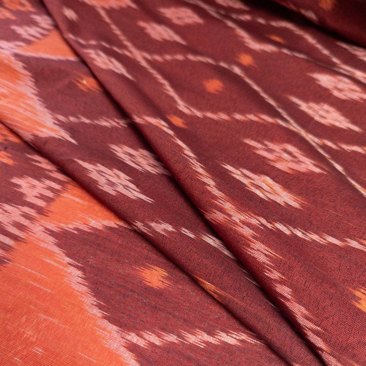 A close-up of folded fabric with an ikat pattern in deep burgundy, rust orange, and light pink, showing the textile's texture and weave.