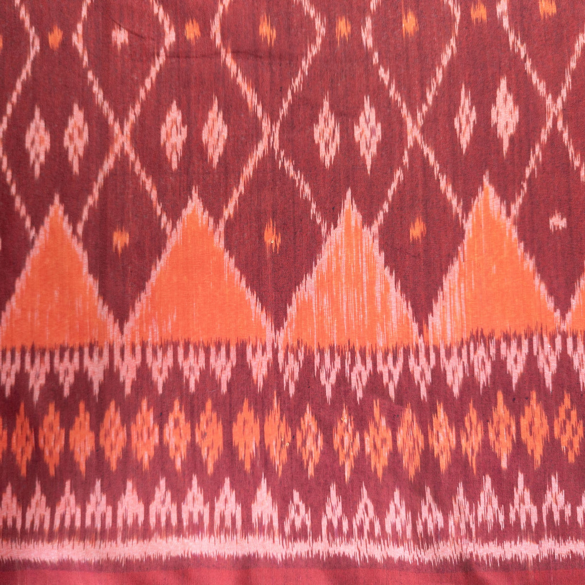 A close-up of a patterned fabric with a deep burgundy background, featuring orange and white ikat-style diamond and triangle motifs.